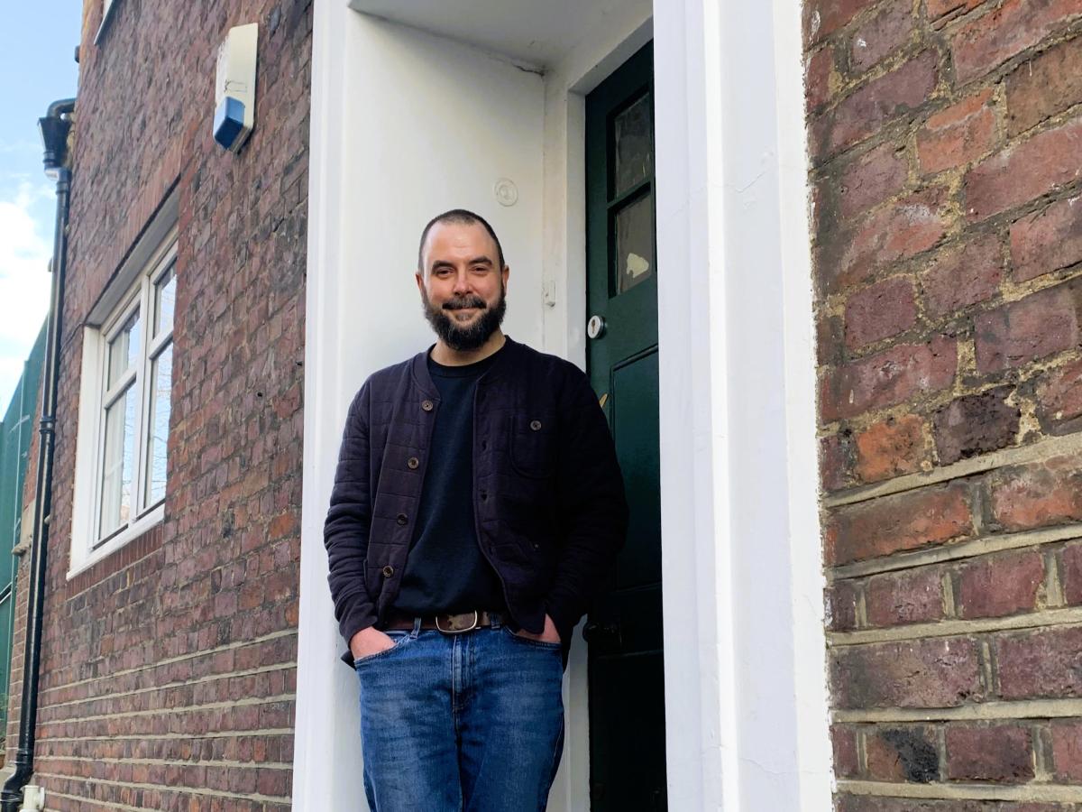 A man stands in a relaxed way on the doorstep of a brick building.