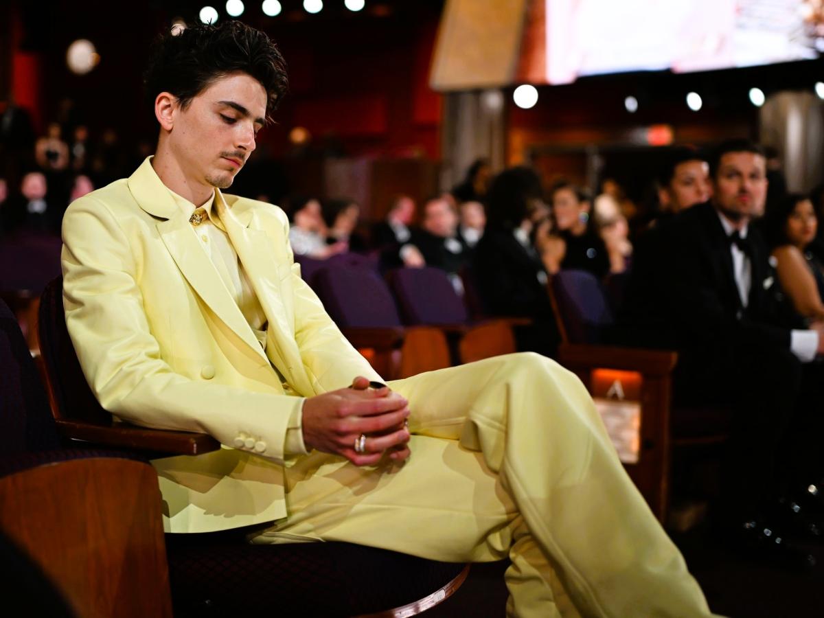 Timothee Chalamet, wearing  light yellow suit reposes in a seat a the Oscars, clasping his hands, his eyes closed.