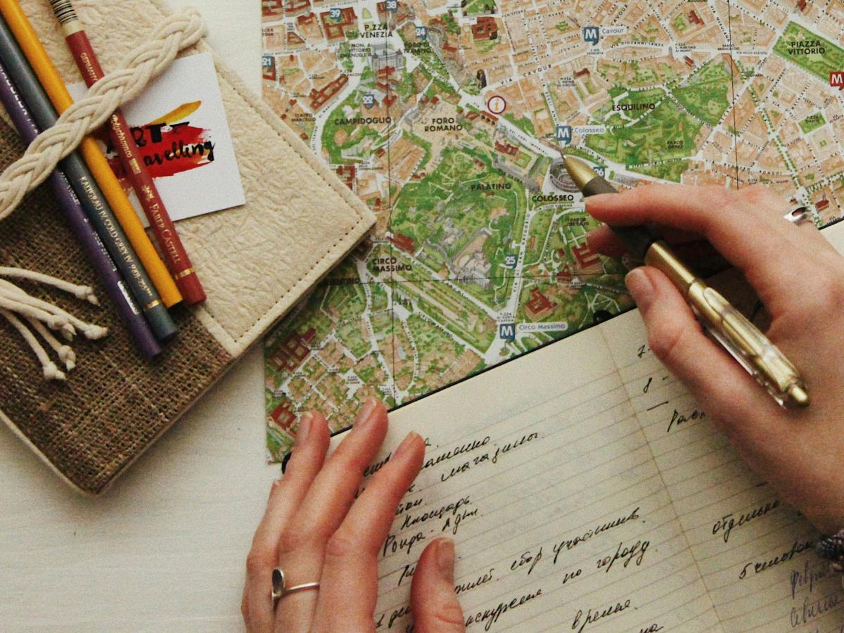 A hand holds a pen over a map, at the side is closed journal and colour pencils.
