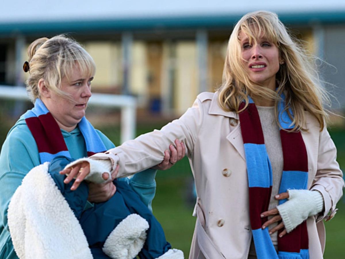 On the sidelines of a pitch a well-dressed mum hands a coat to a sceptical looking mum beside her.