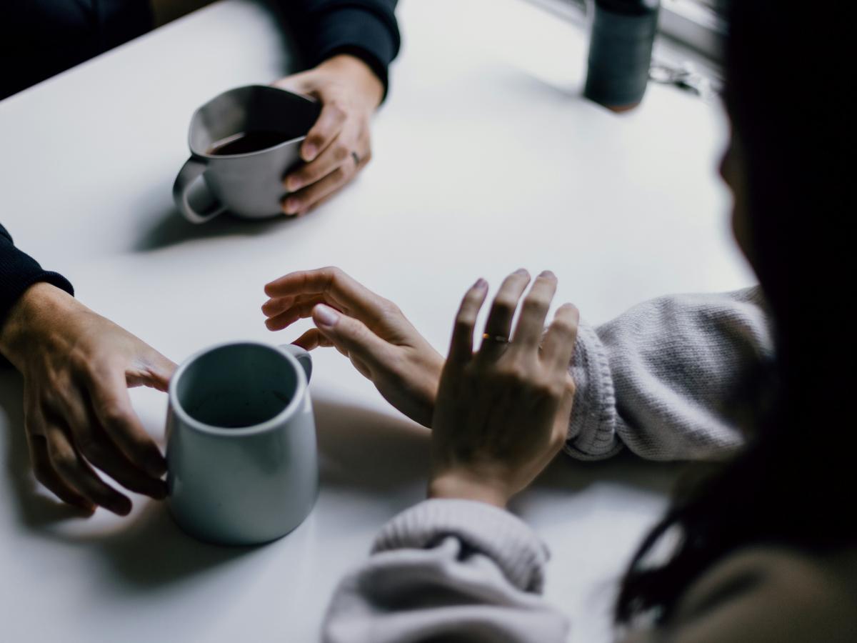 A cup of coffee is offered across a table to a hand that is hesitant.