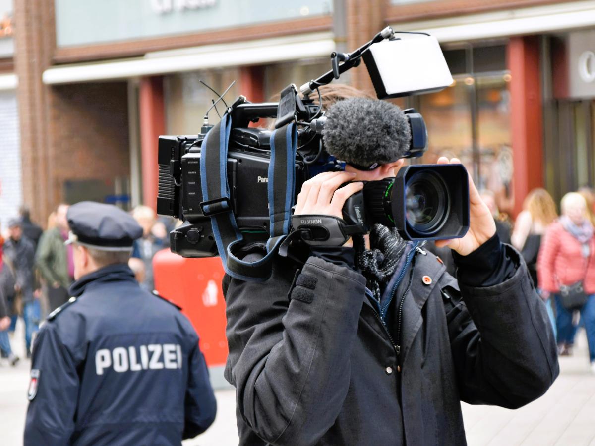 A news cameraman holding a camera, stands back to back to a police officer.