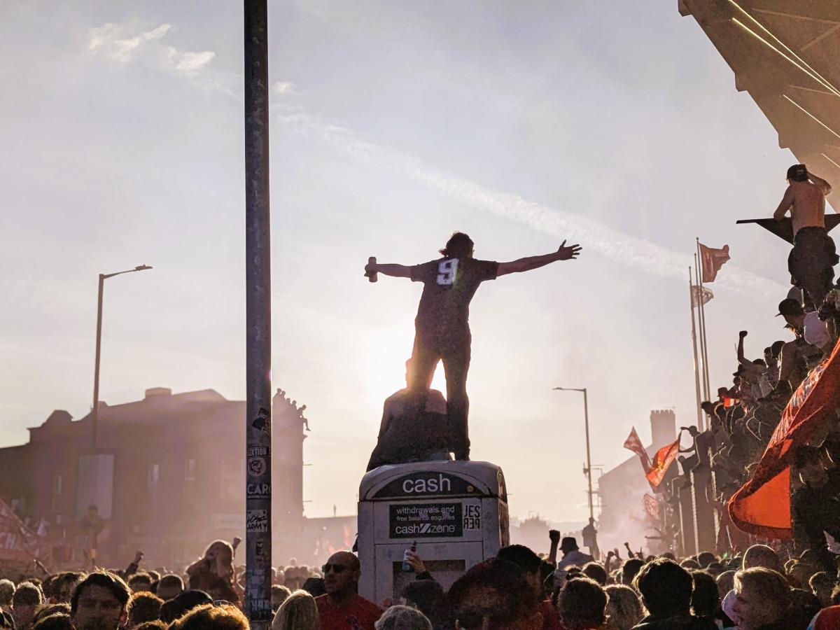 Amid celebrating football fans, one stands on top of a kiosk with outstretched arms.