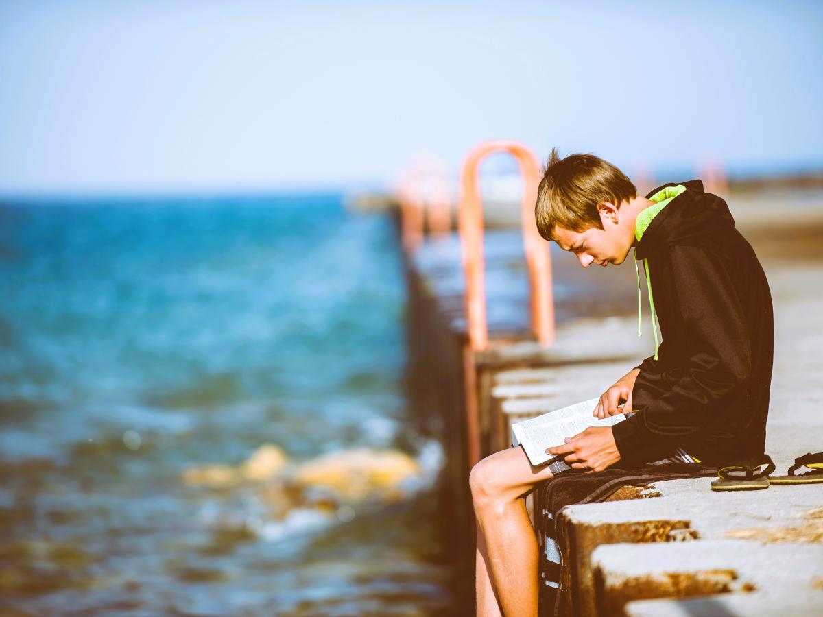 A man sits on a pier intently reading a book on his lap.