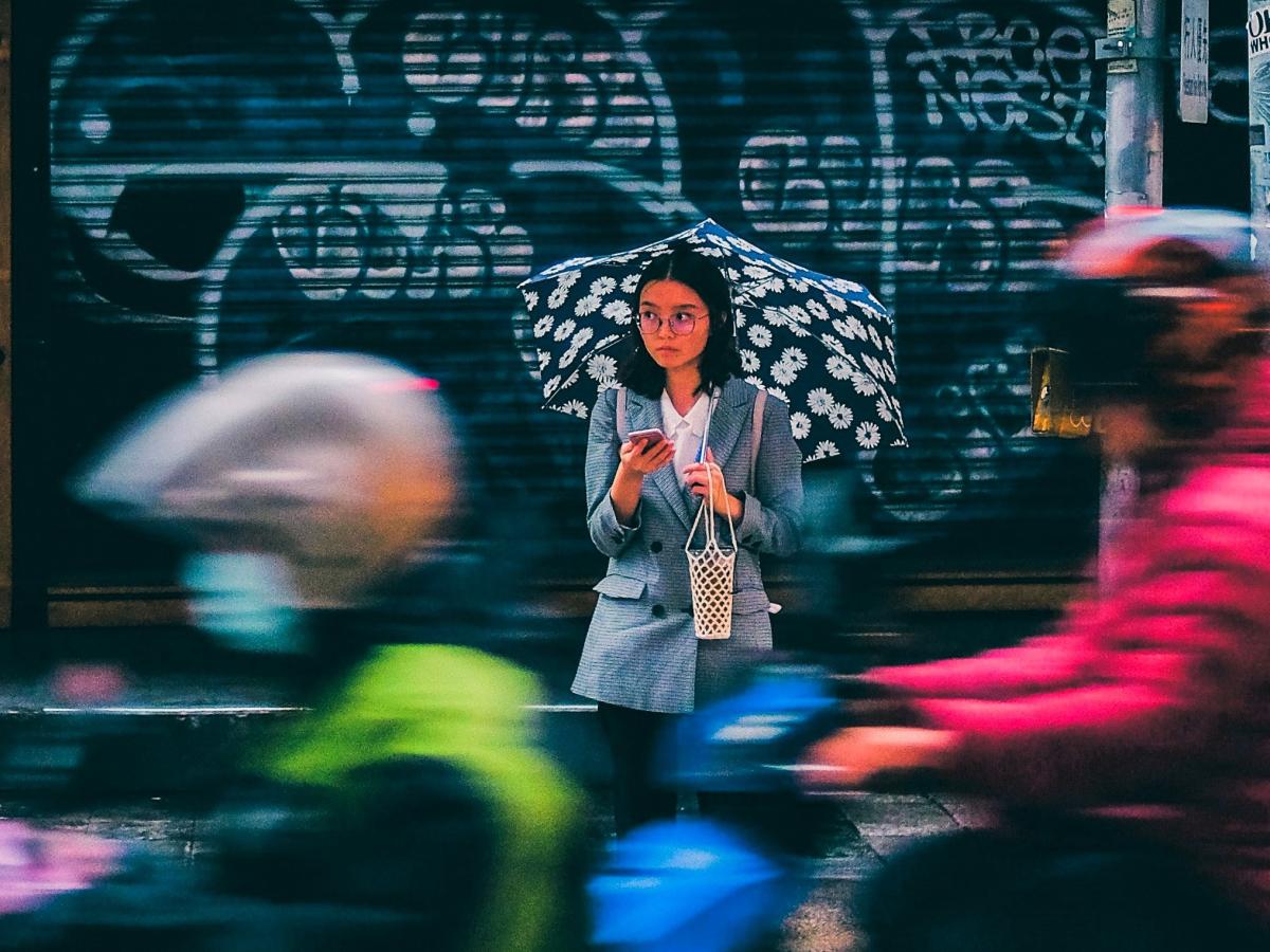 A woman stands across a busy roads, looking up from her phone in a sad way.