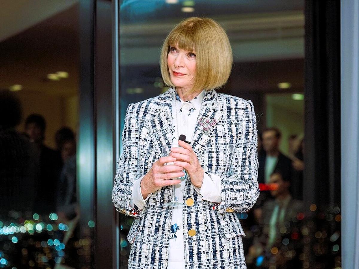 Anna Wintour stands holding a small mic.