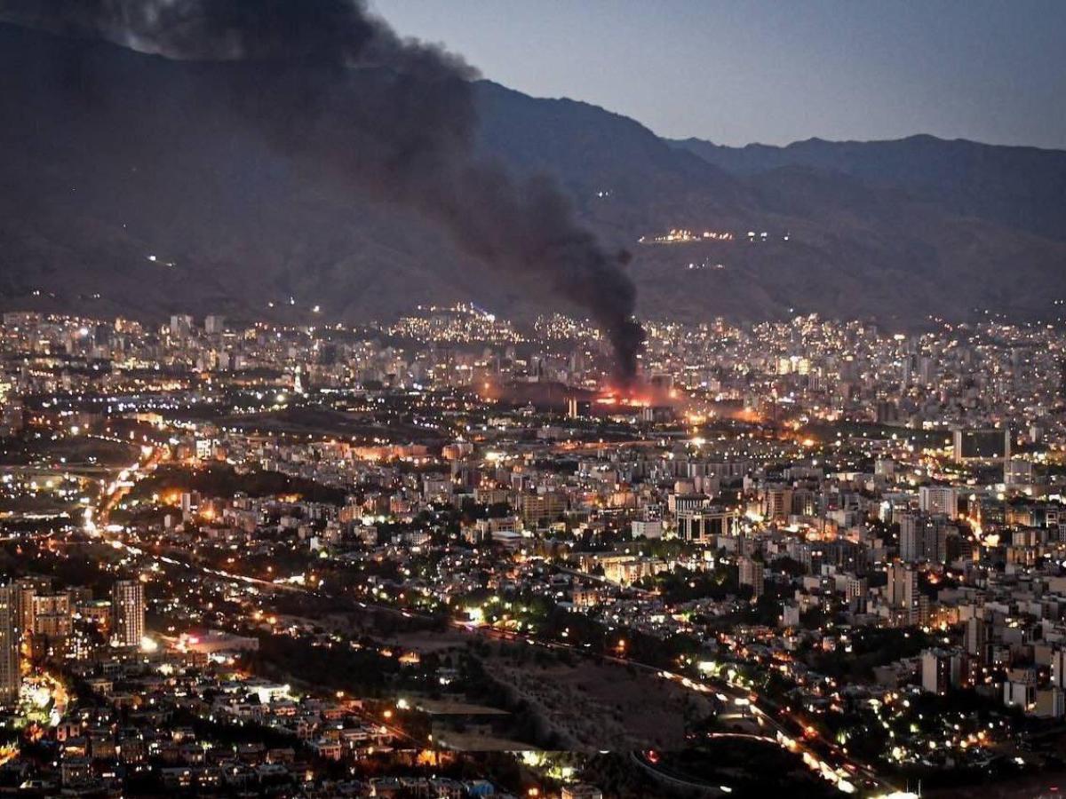 Smoke rises from the site of a bomb amid a high wide view of a city at twlight
