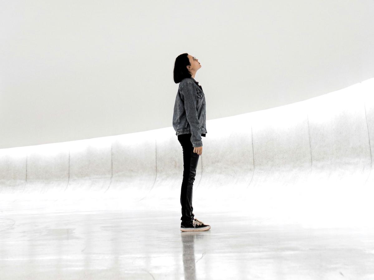 A woman stands against a sparse white background looking up.
