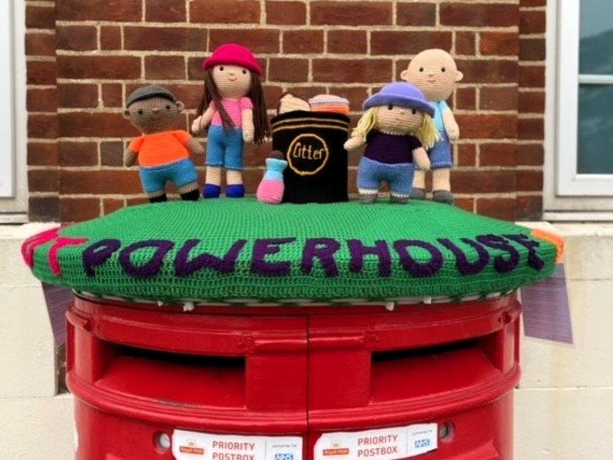 A knitted post box topper shows a group of people and the word powerhouse.
