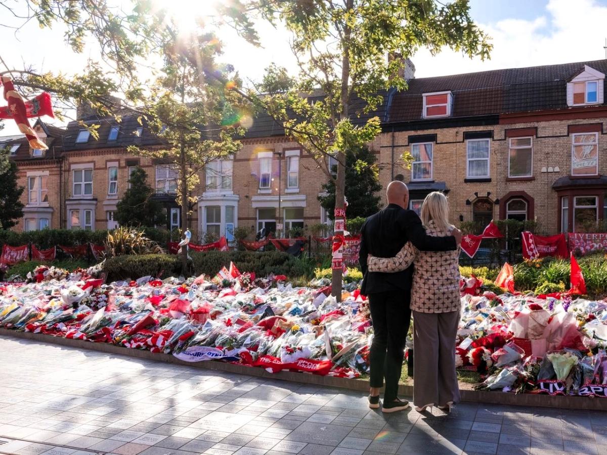 A couple hold each other as they look at floral tributes on the ground