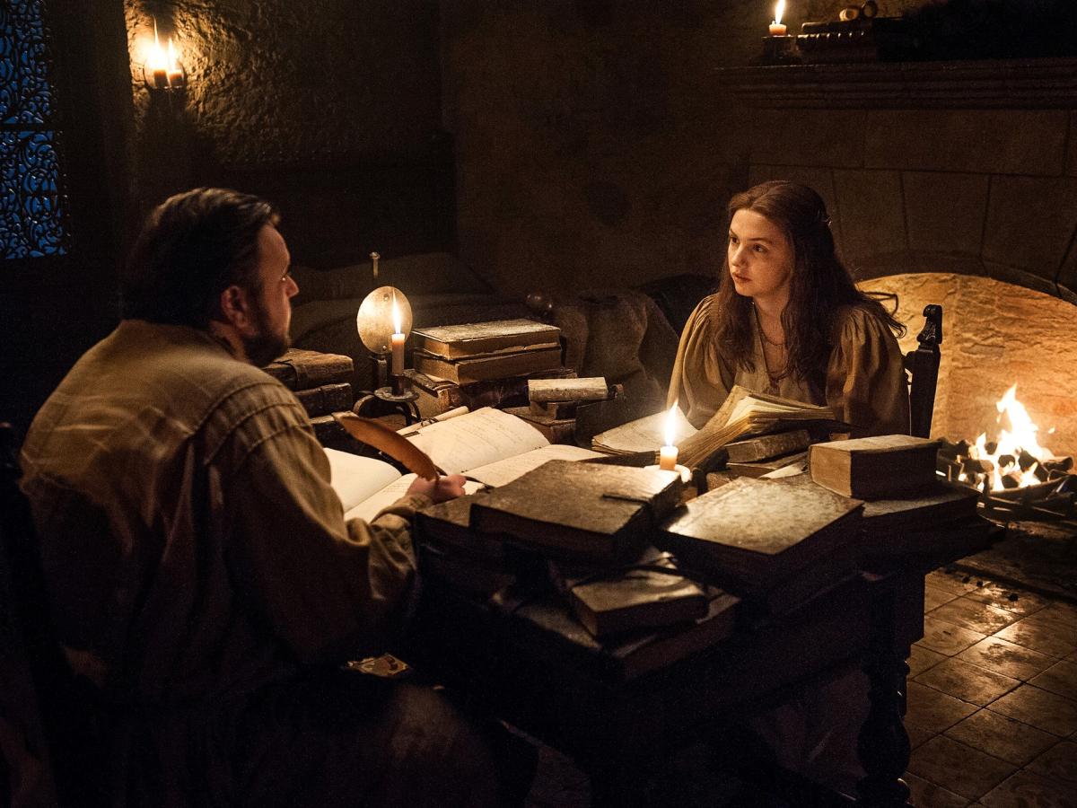 Two people sit at a table strewen with old books lit by candle light.