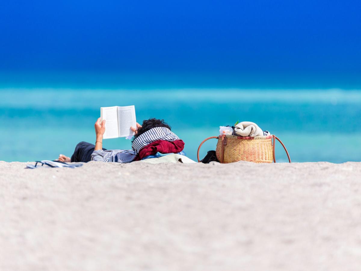 A person lying on a beach holds a book up to read.