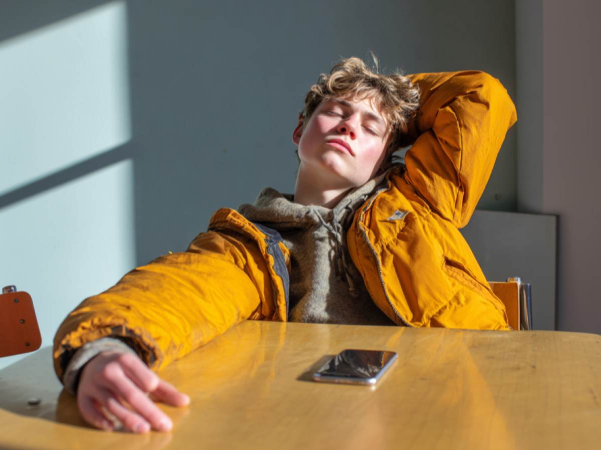 A seated teenager stretches back bored, a phone is on the table in front of them