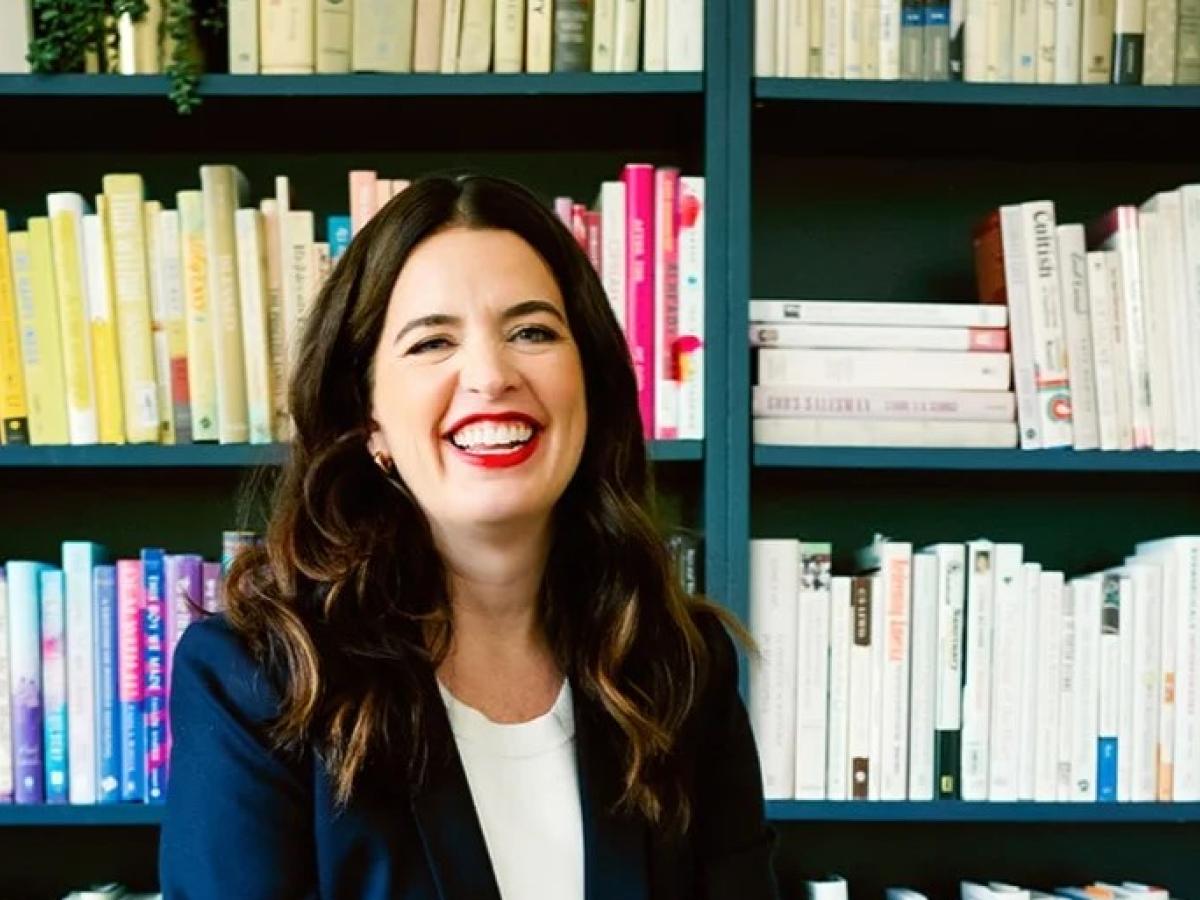 A woman sits and smiles in front of her bookcase.