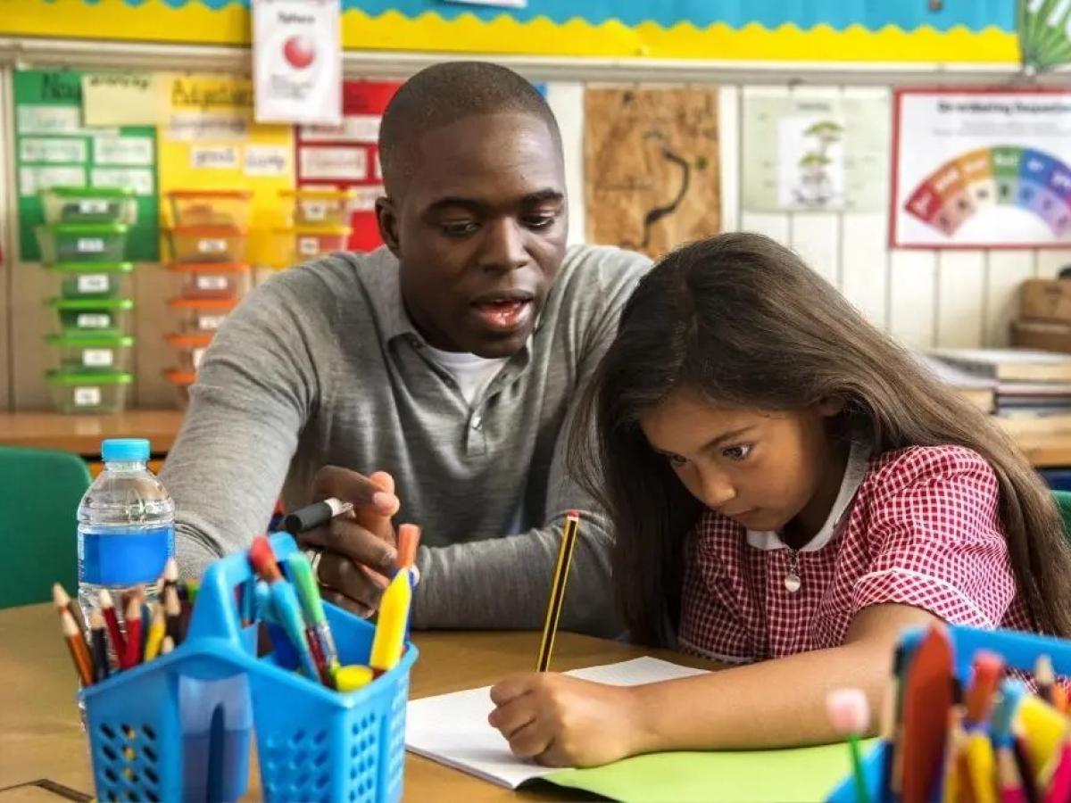 A teacher looks on as a young child concentrates on writing.