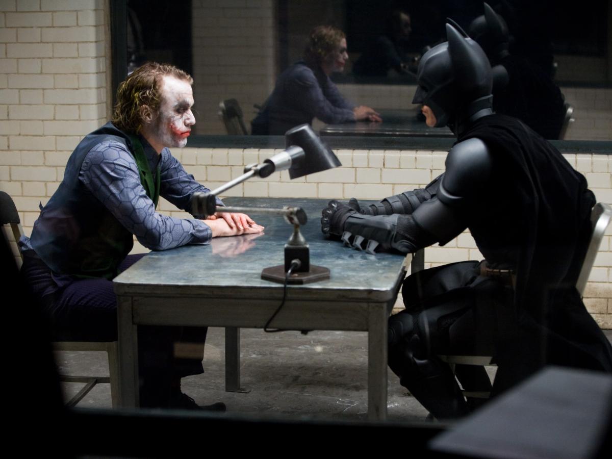 The Joker and Batman sit opposite each other in an interrogation room.