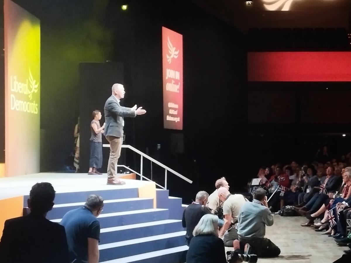 Tim Farron stands on a conference stage with arms wide