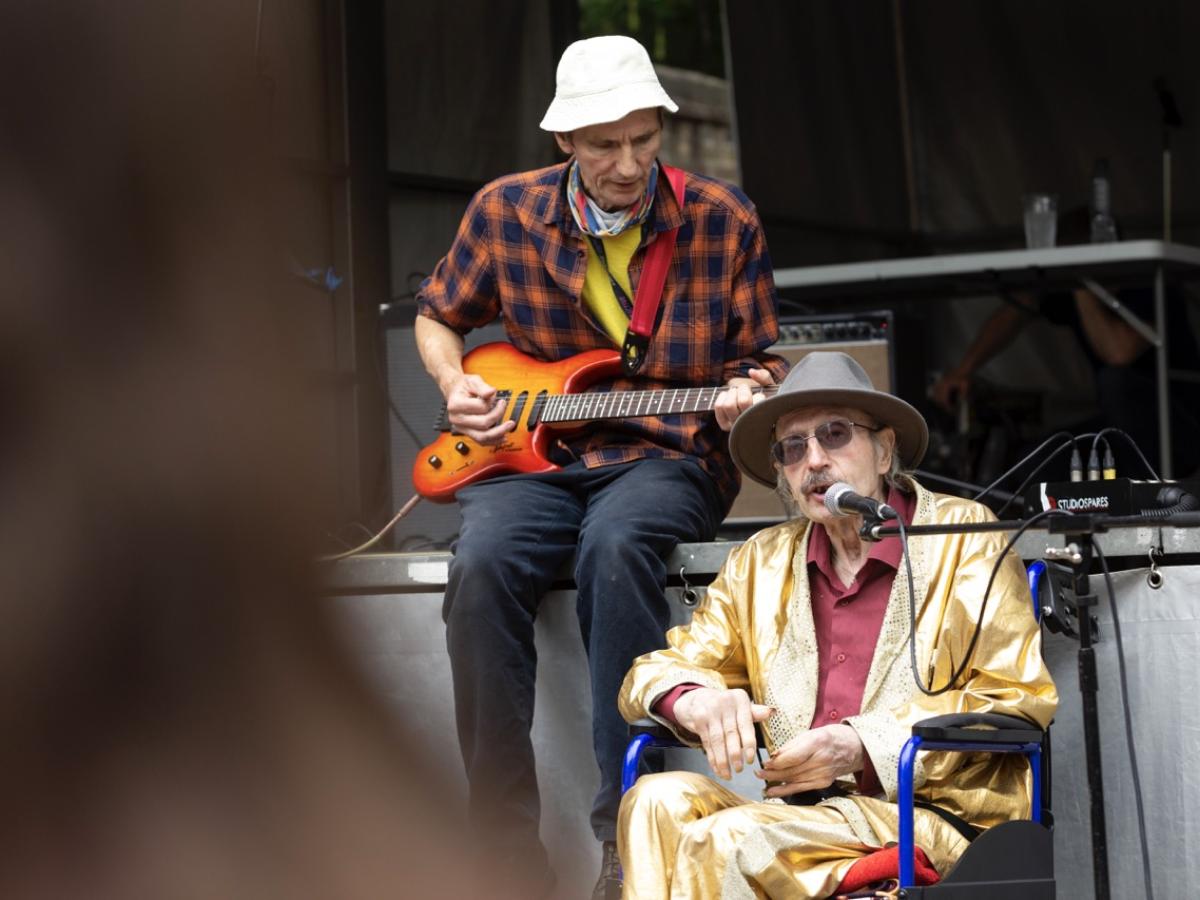 An elderly Elvis impersonator sits on a stage wearing a gold suit.