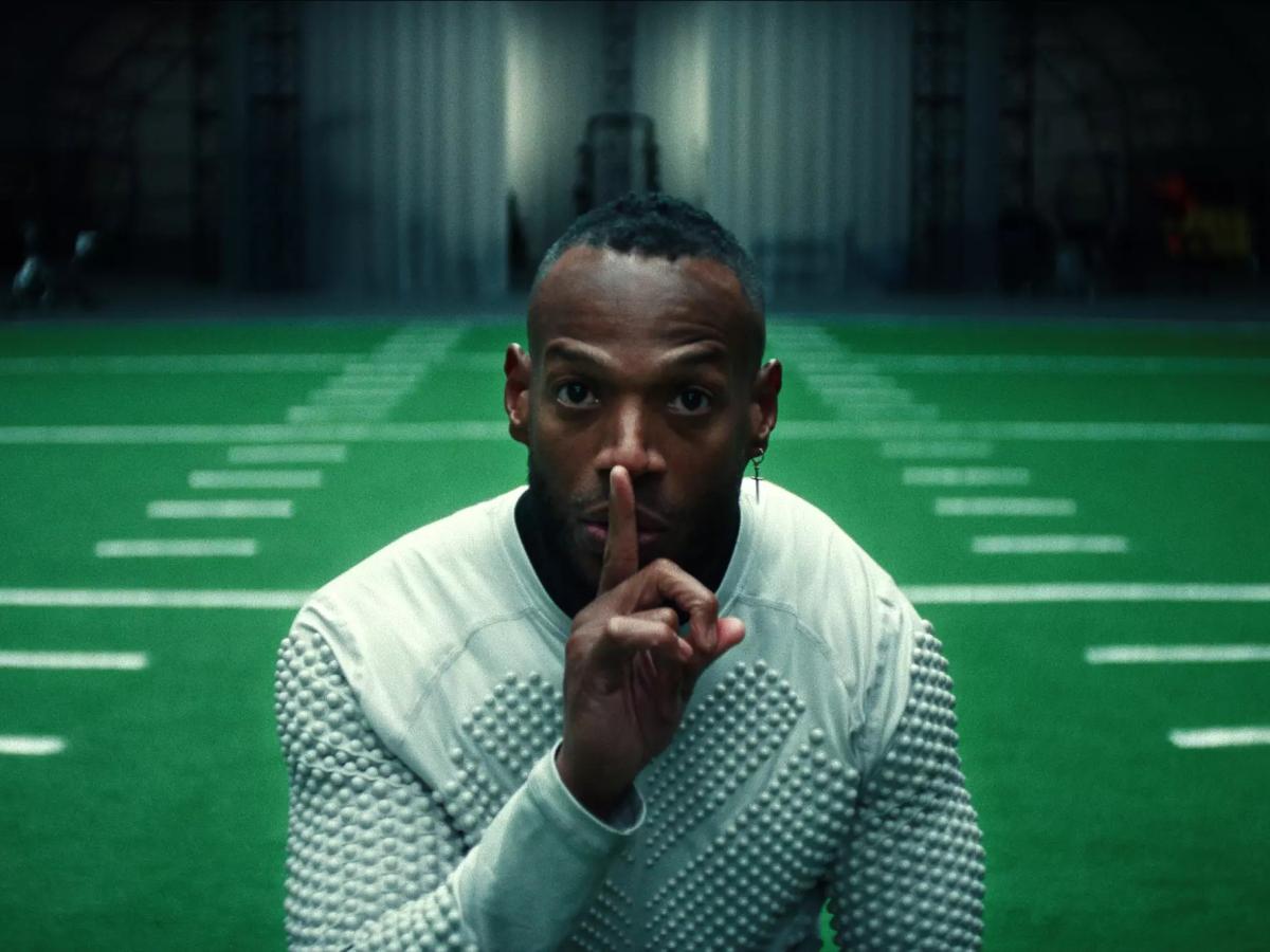 A player holding a finger to his lips stands in front of an indoor American football pitch.