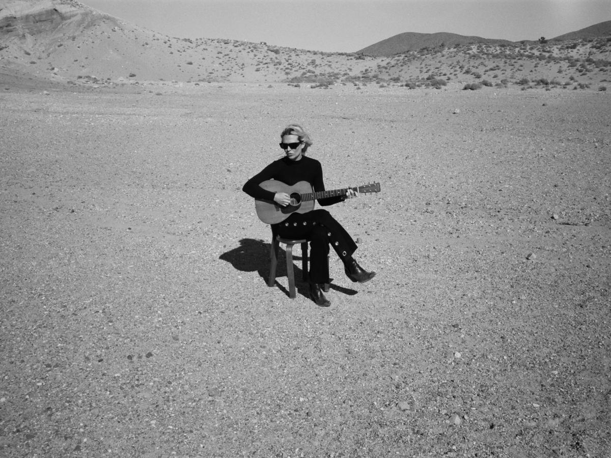 A musician wearing black sits on a chair in a desert holding her guitar.