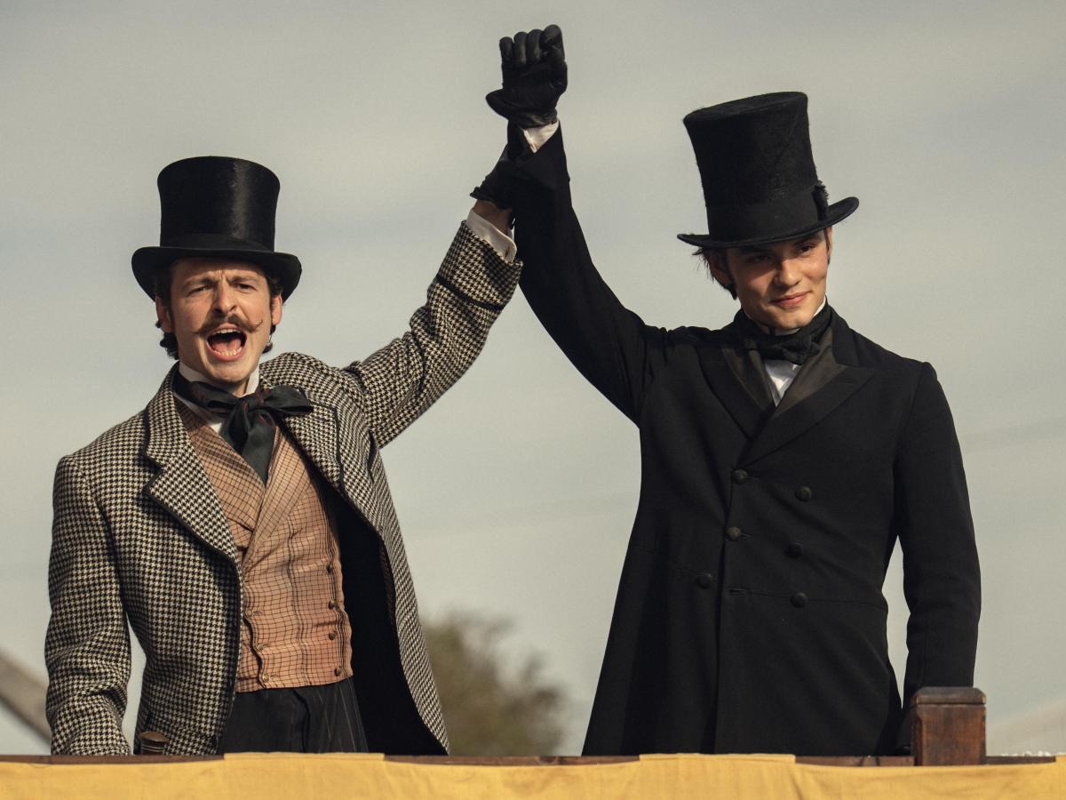 Dressed in Victorian clothes, two brothers raise their arms together.
