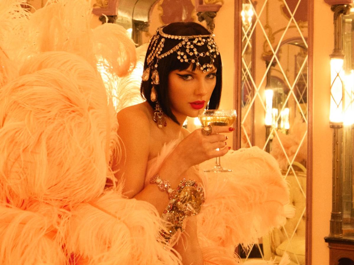Taylor Swift, dressed as a showgirl, sips from a glass.