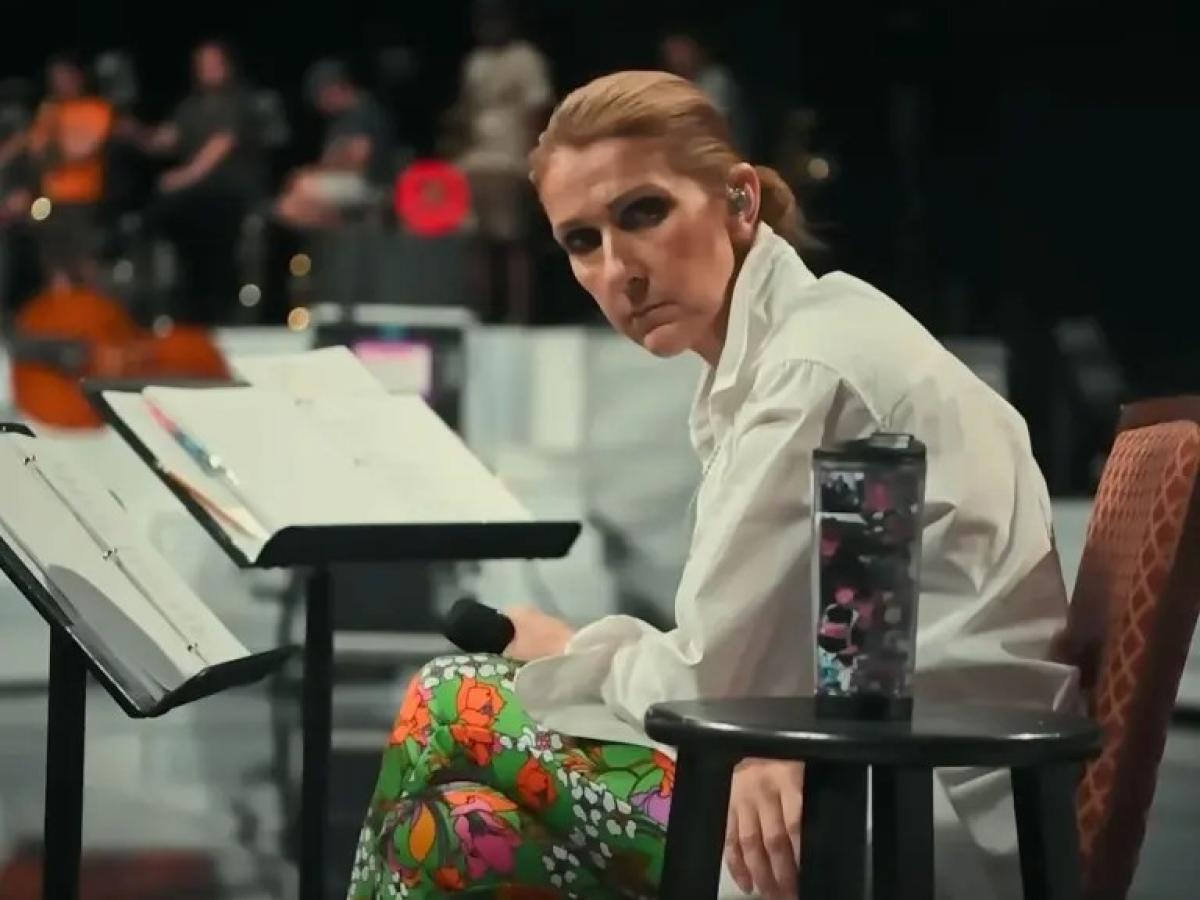 A seated Celine Dion, leans forward, head to the side, holding a mic.