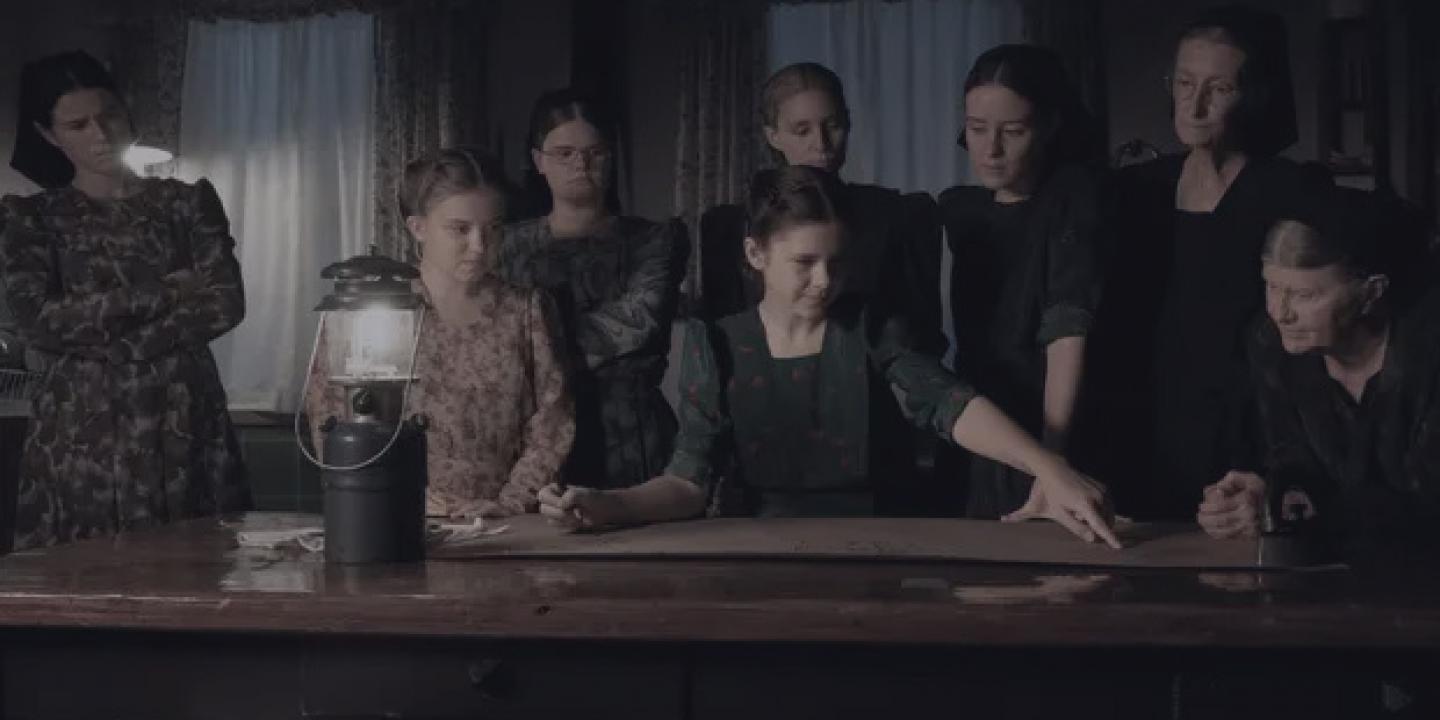 a group of women stand and sit around a table lit by a gas lamp.