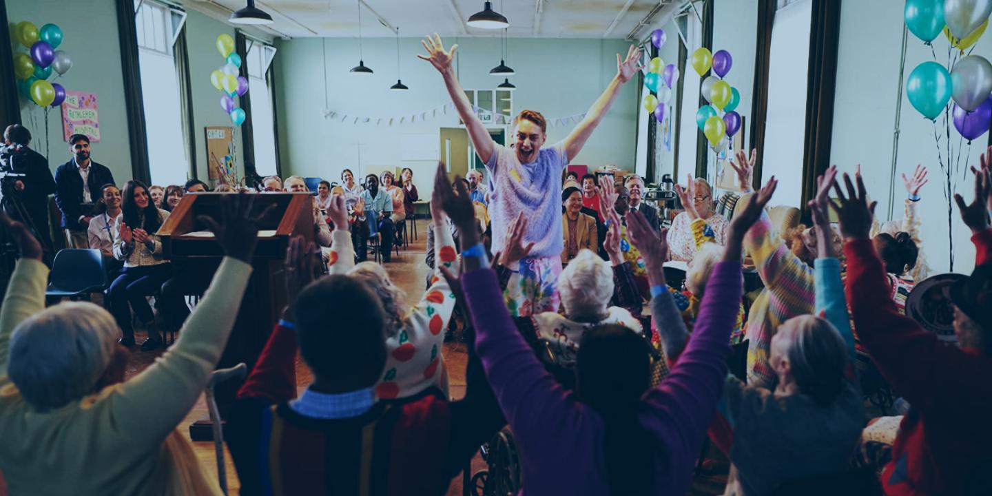In a hall decorated for a celebration a person stands in front of a seated group, all have their arms raised in celebration.