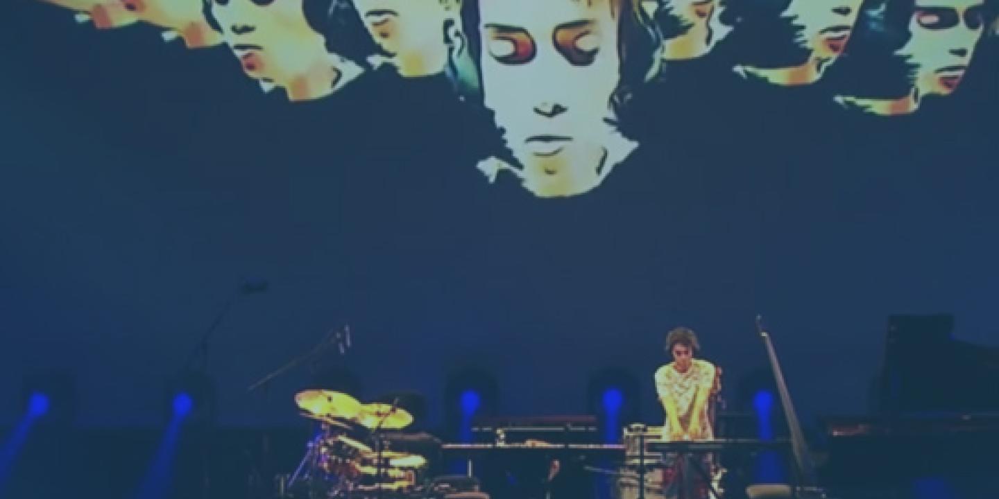A muscian plays a keyboard on a concert stage surrounded by instruments, while multiple images of his face are projected behind him.