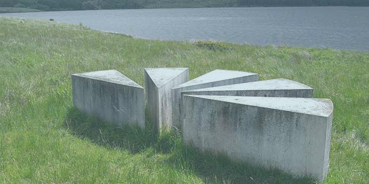 A set of low concrere blocks in the shape of a map's viewpoint symbol sit beside a lake.