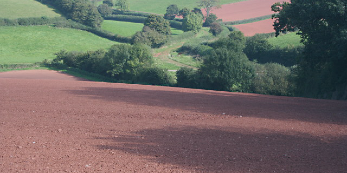 A ploughed field of red soil is in the foreground, sloping down into a valley with a track and green fields beyond