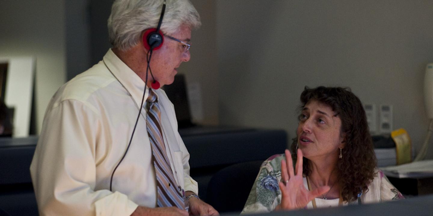 Two scientists talk to each other at console in a space mission control room.