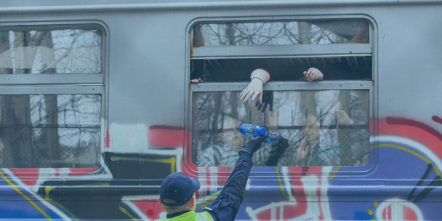 A helper in a yellow vest reaches up to a open train carraige window while offering a bottle. The side of the carraige is covered in graffiti.