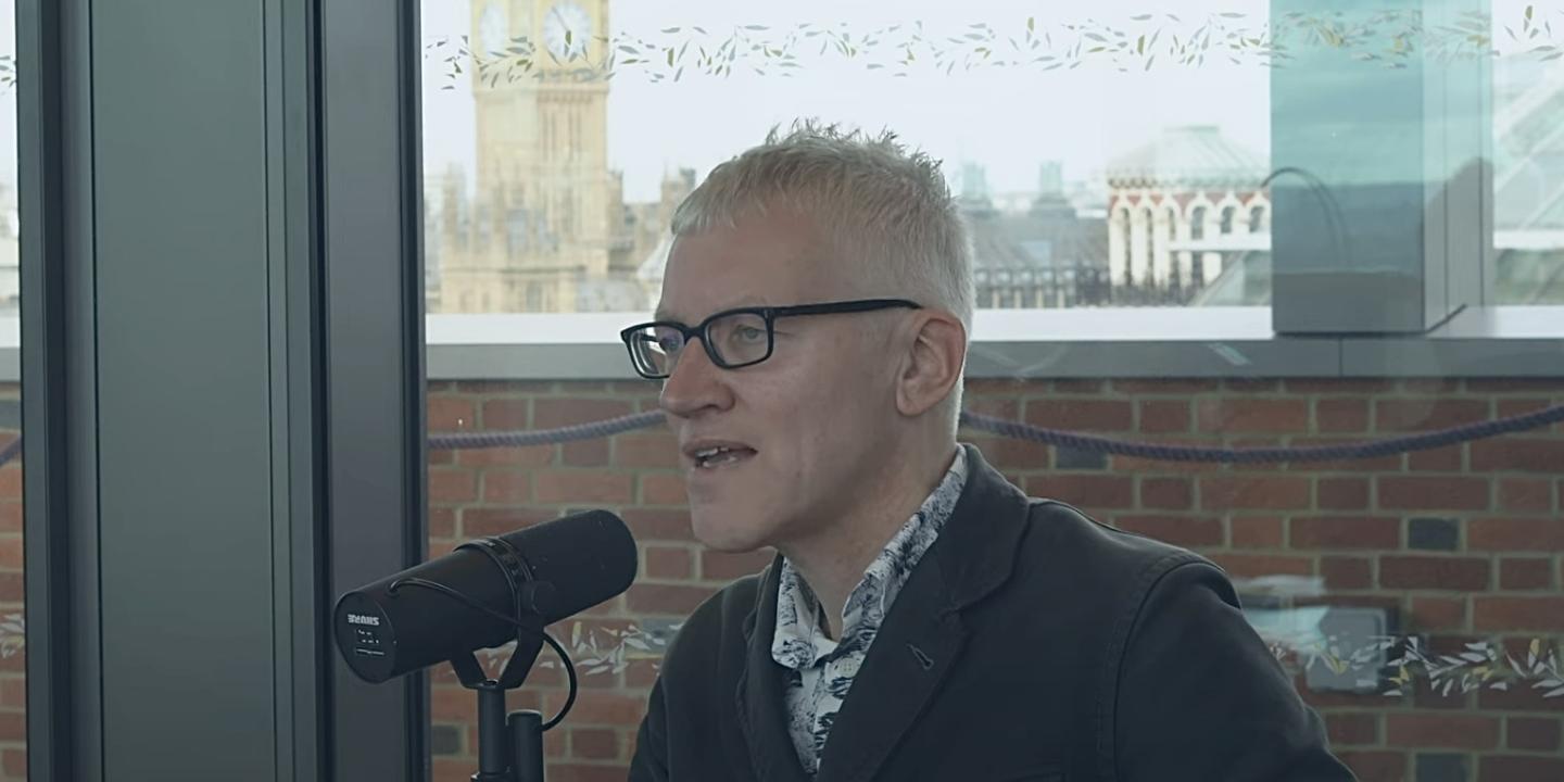 A man sits at a table speaking into a microphone but looks into the distance. Behind him, through the window and beyond a wall is Big Ben