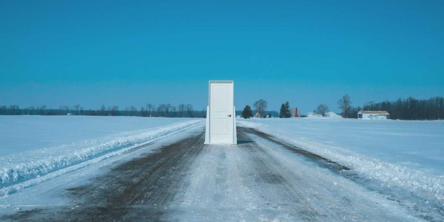 A theatrical set door stands in the middle of a snowploughed road between fields of snow under a blue sky.