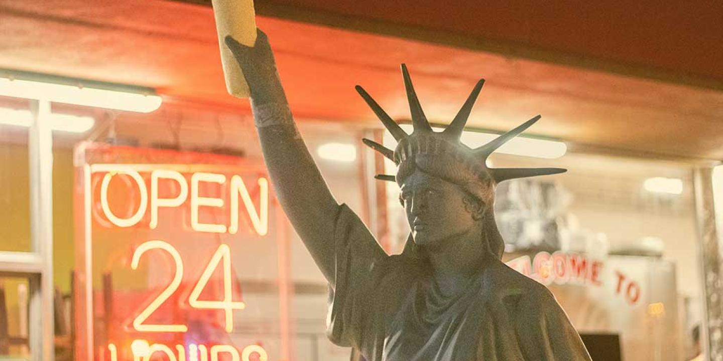 A copy of the Statue of Liberty, holding a stick of bread, stands outside a shop window displaying an 'Open 24 Hours' signs.