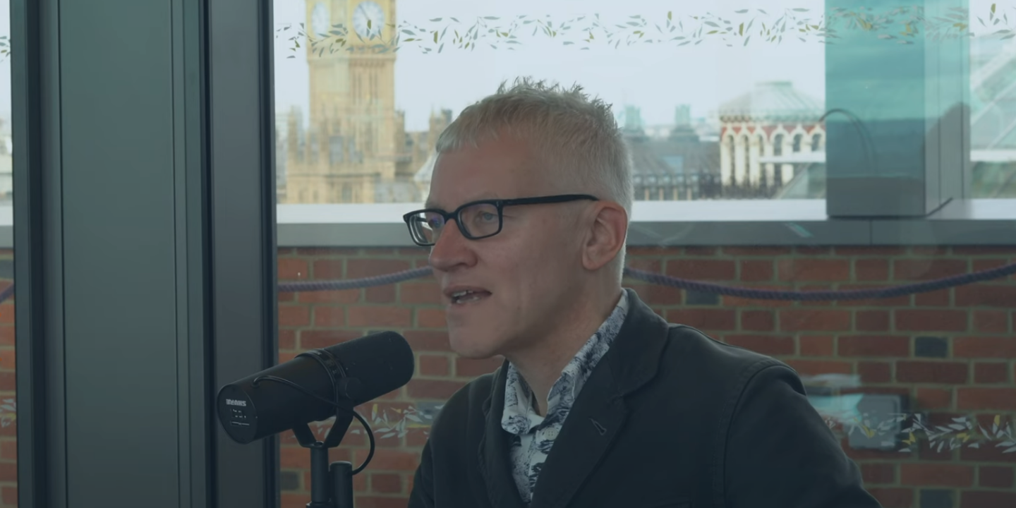 A man sits at a table speaking into a microphone but looks into the distance. Behind him, through the window and beyond a wall is Big Ben