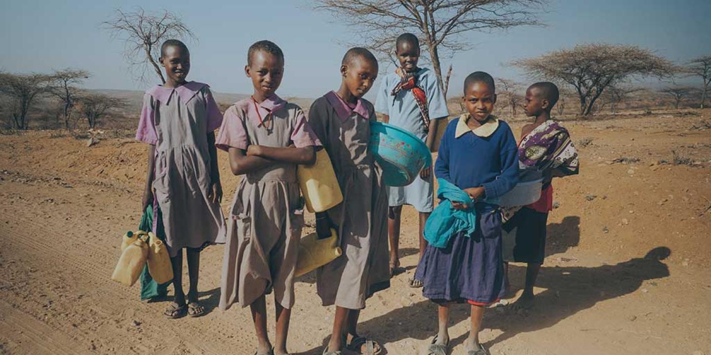 Girls stand in the northern Kenyan scrubland holding water bottles.