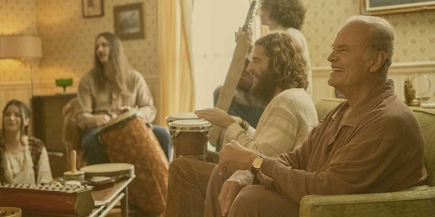 A group sit in a lounge playing musical instruments while the man closest to the camera laughs.