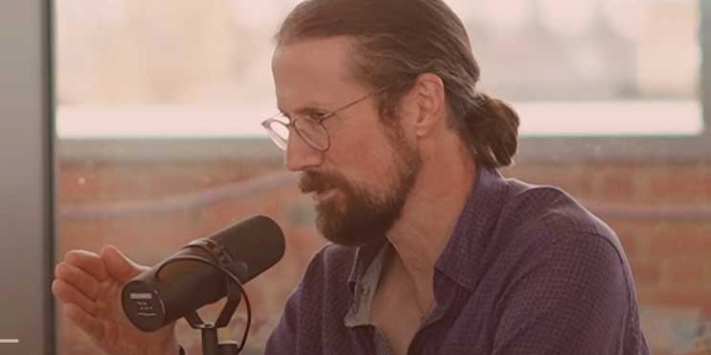 A sitting man speaks into a microphone will gesturing with one hand