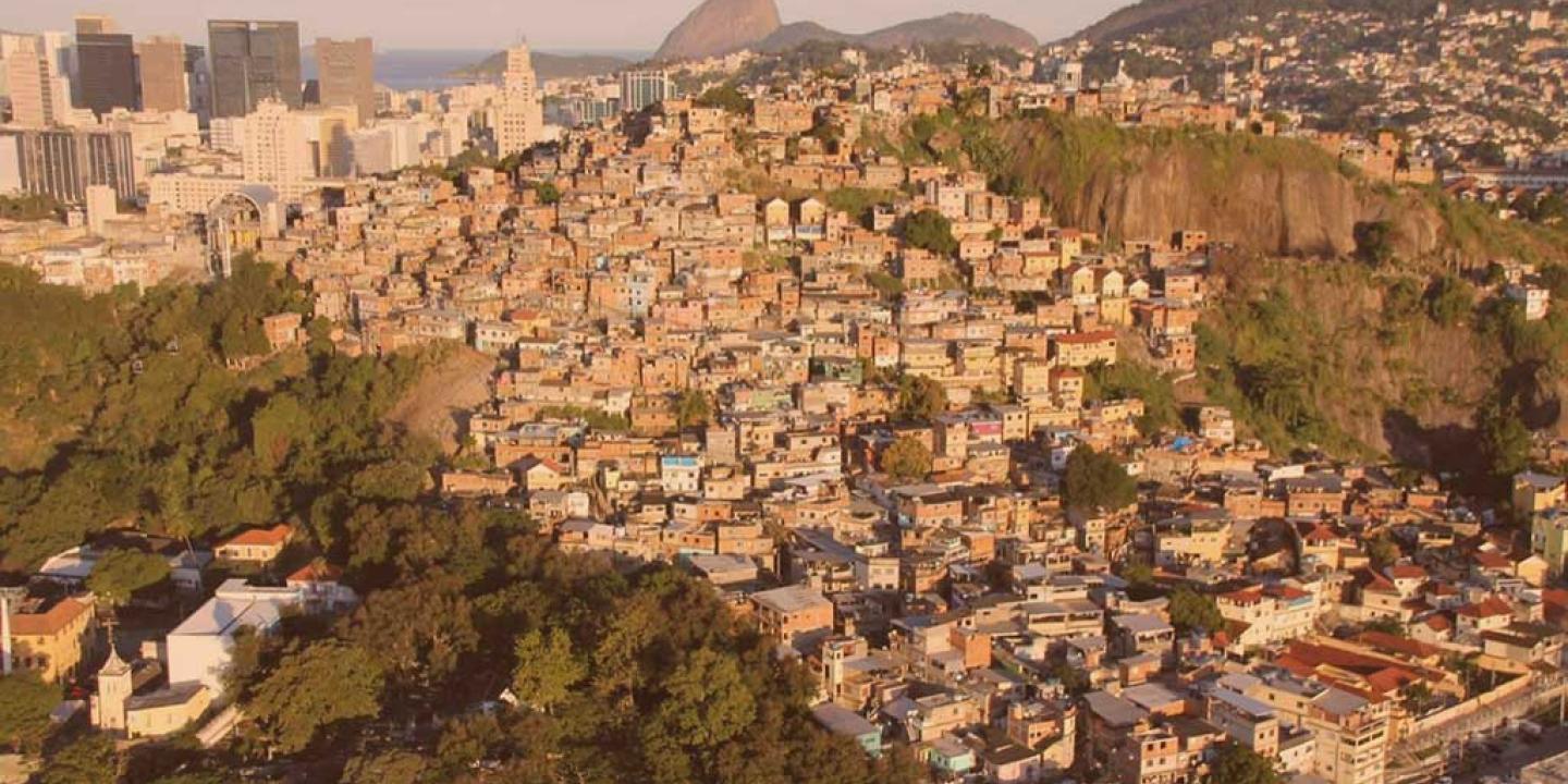 an aerial view of a shantytown on a steep hill side in Rio, Sugerloaf Mountain is visible in the distance 