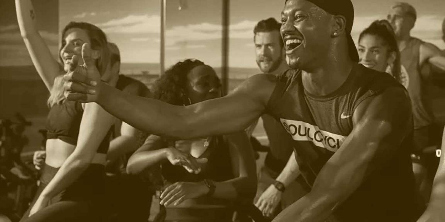 A spin class rider smiles and gives the thumbs up sign in front of other riders.