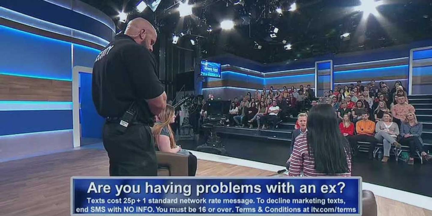 A view across a talk show TV set showing a security guard standing between two guest while the host talks to them. An audience looks on.