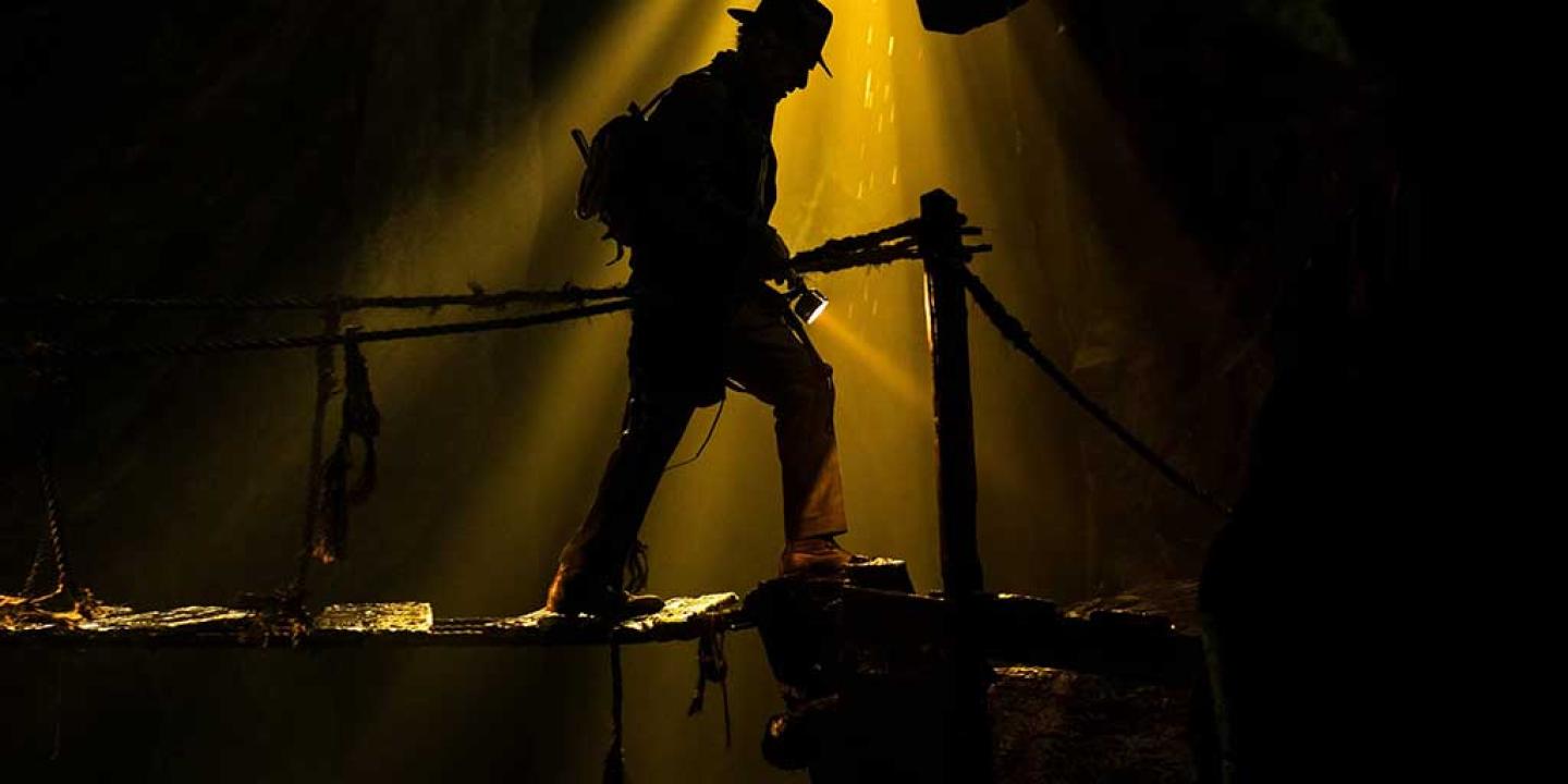 A silouhette of an adventurer, wearing a fedora hat, stepping gingerly along a rickety bridge.
