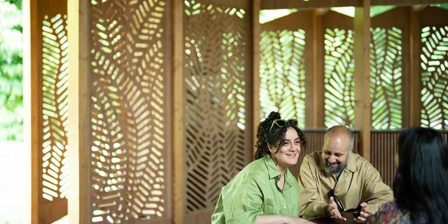 Around a table, against a backdrop of fret-cut wood, three people talk and listen to each other with great interest.