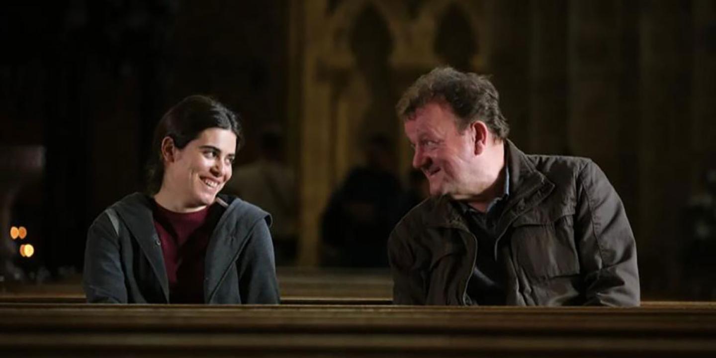 A man and a woman sit in a cathedral pew and incline their heads towards each other.