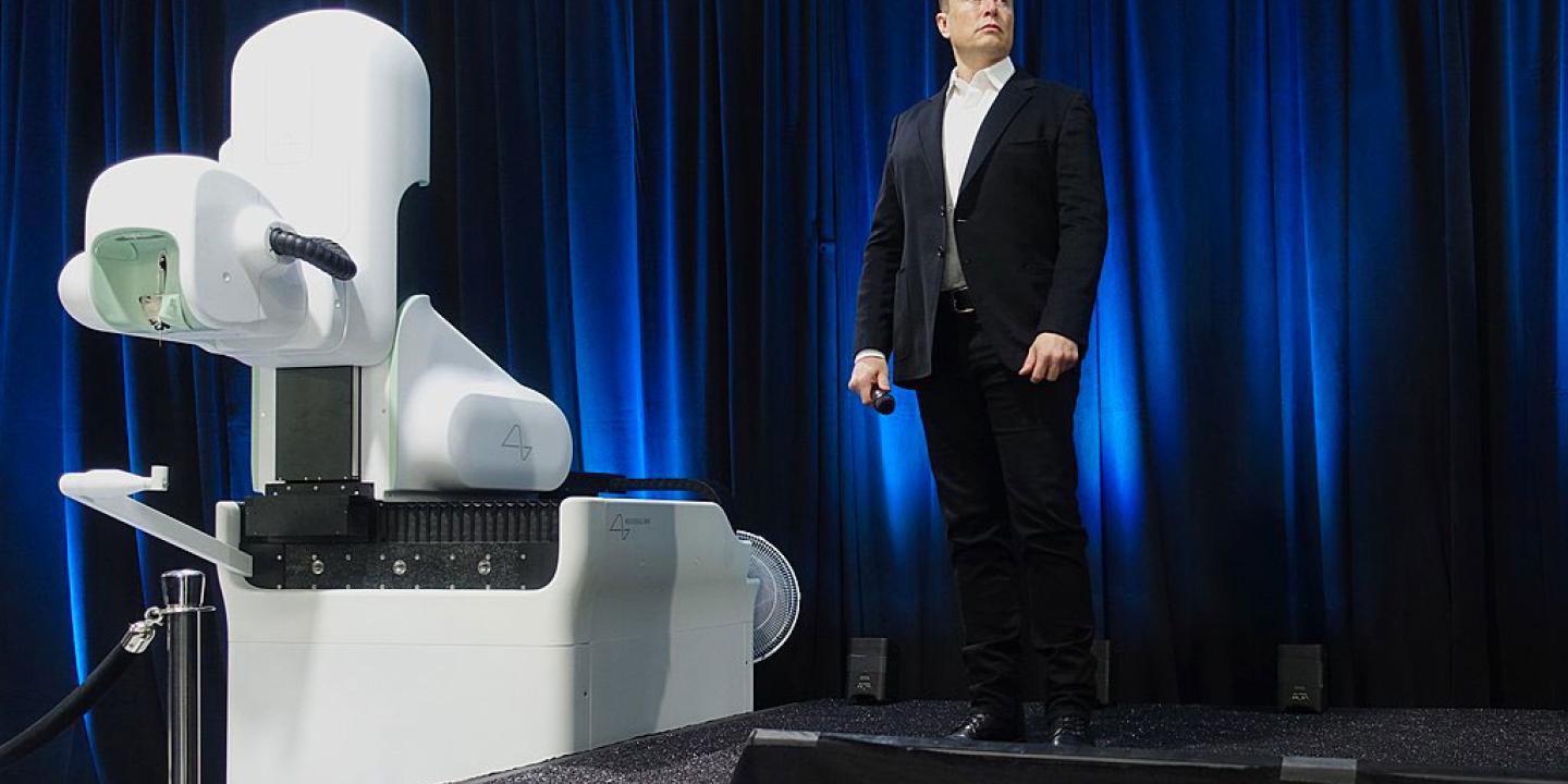 Elon Musk, wearing a dark suit, stands on a stage to a white robotic looking surgical robot.