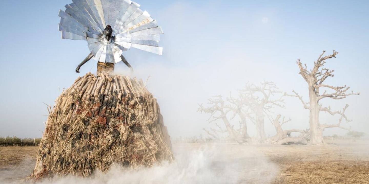 A photo-realistic work of art depicting a fiigure raised on fire wood wearing a colour of large metal rays, against a desertifying landscape of dead trees.