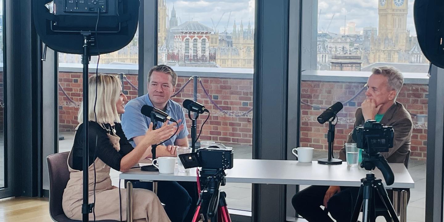 Around a table with microphones, three people record a podcast, one leans in talking and gesturing with a hand while the others listen.